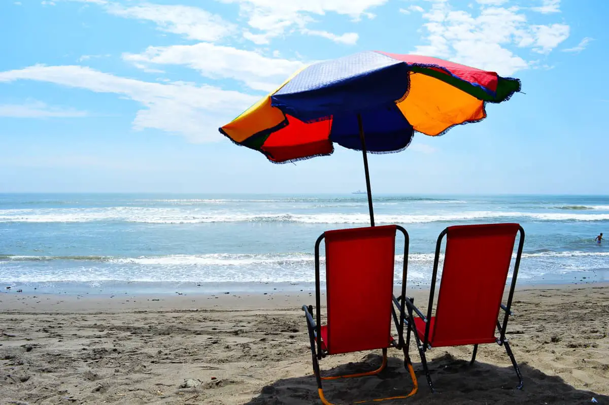 Two red chairs and a colorful umbrella at the beach looking towards the sea. Beautiful blue sky with a bit of sun.