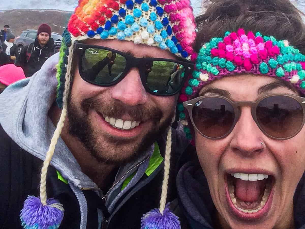 Young man and woman in the snowy mountains. Close up of their faces, they are both laughing and excited. Both wear Peruvian hats in traditional colors and patterns. Both wear sunglasses.
