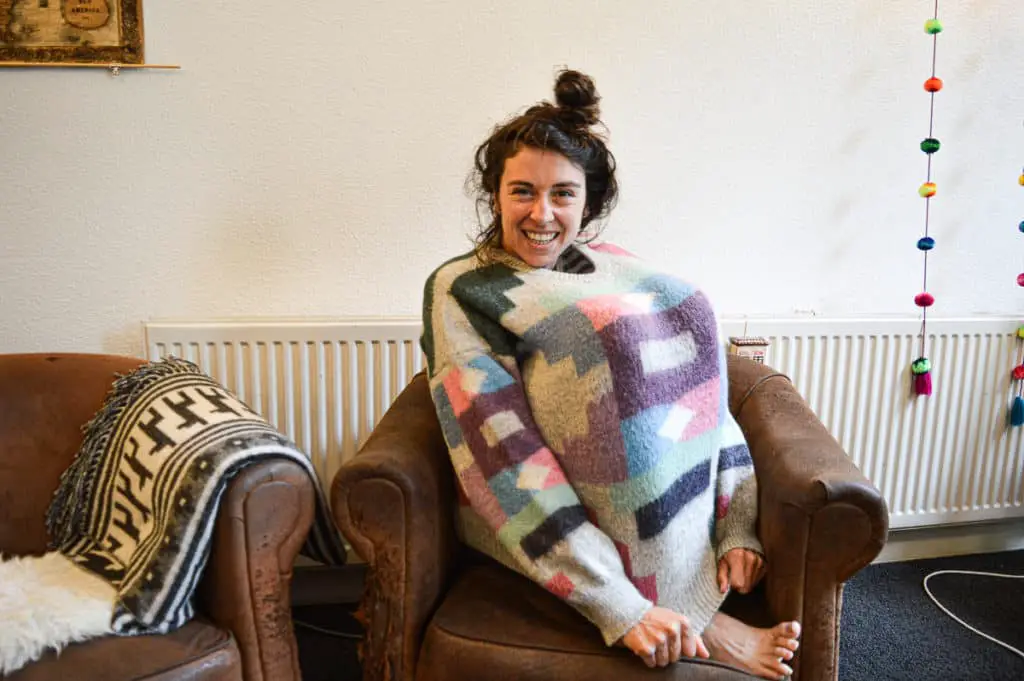 Young woman (Eveline) sitting on a chair with her knees pulled in and a sweater wrapped around them. The sweater is made of alpaca wool and looks very stretchy.