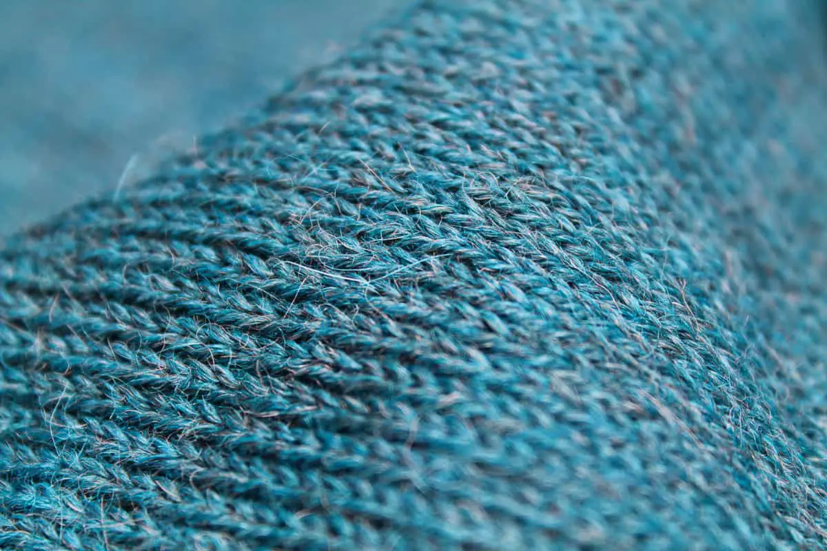 100% alpaca woolen scarf. Knitted scarf with medium fine sitch. Crocheted border and vegan "leather" label. Blue melange with tones of green and grey. Close up of stitch and colors.